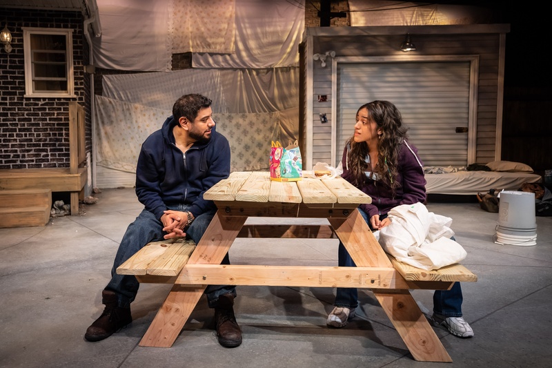  Two people sitting at a wooden picnic table on a stage set. 