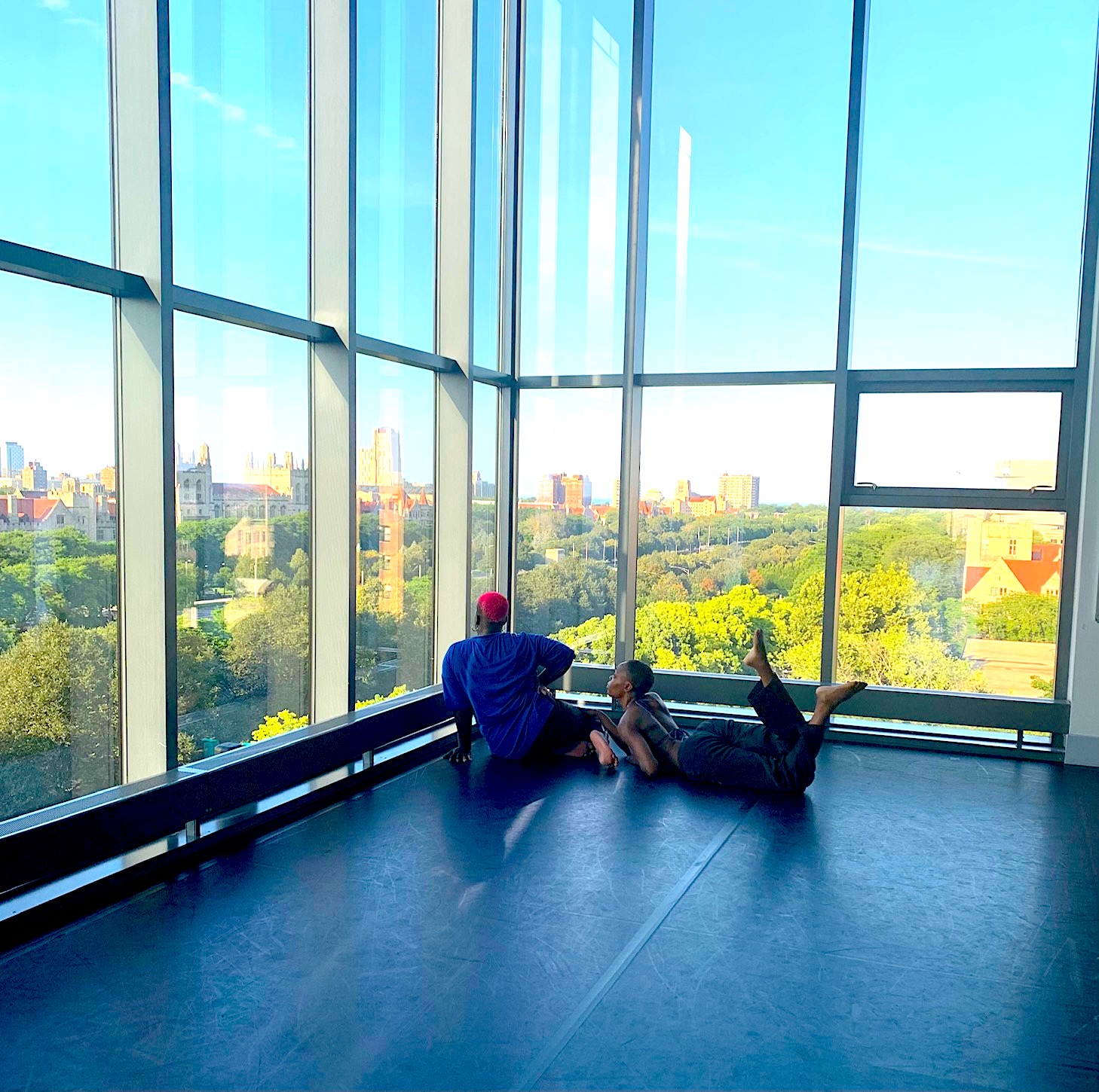  Two dark-skinned Black dancers improvise in front of floor to ceiling windows showcasing the Chicago skyline 