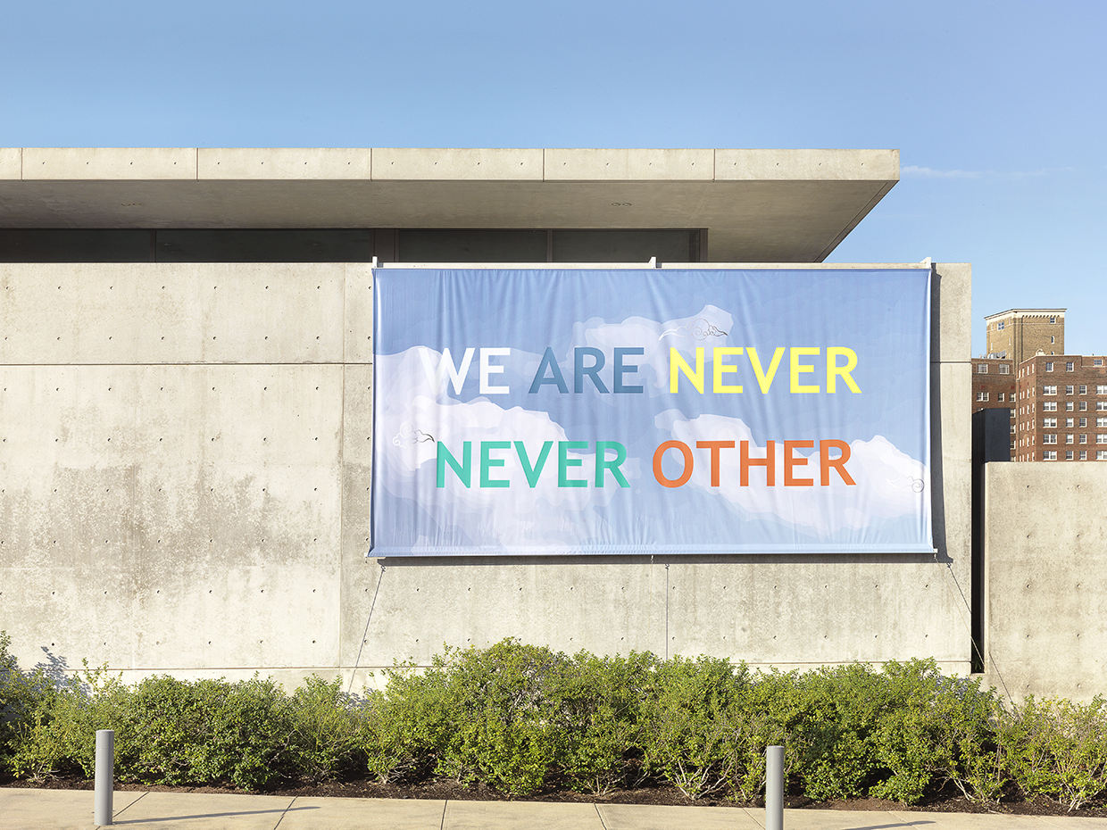 A large vinyl banner hangs on the facade of a large concrete building. The banner is blue with clouds and there is colorful text that reads We Are Never Never Other.