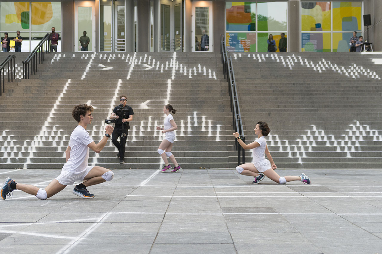 Three dancers in white shirts and shorts with white knee pads moving in athletic lunging positions across the paved plaza in front of the Museum of Contemporary Art Chicago, surfaces marked with a white chalk pattern and camera man chasing the dancers.