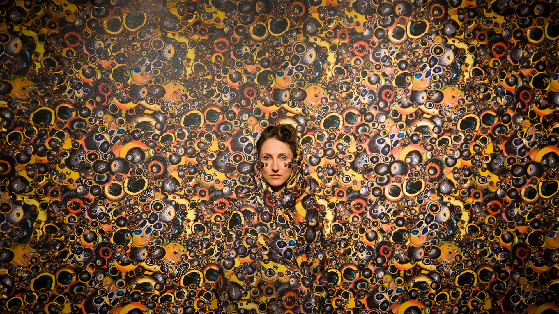 A person draped in a orange, yellow, and black patterned fabric, standing in front of a wall covered in the same pattern.
