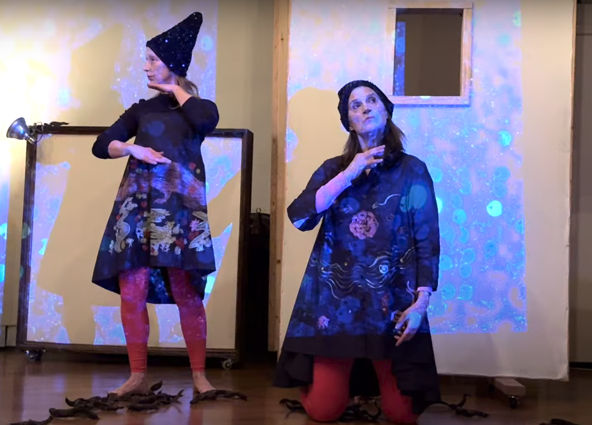 Sara Zalek and Elaine Lemieux in front of projected blue sparkling light against a mix of white walls. Sara is in profile looking left, Elaine faces more frontal, her gaze looking upward right. They both are mid gesture with strong concentrated look.