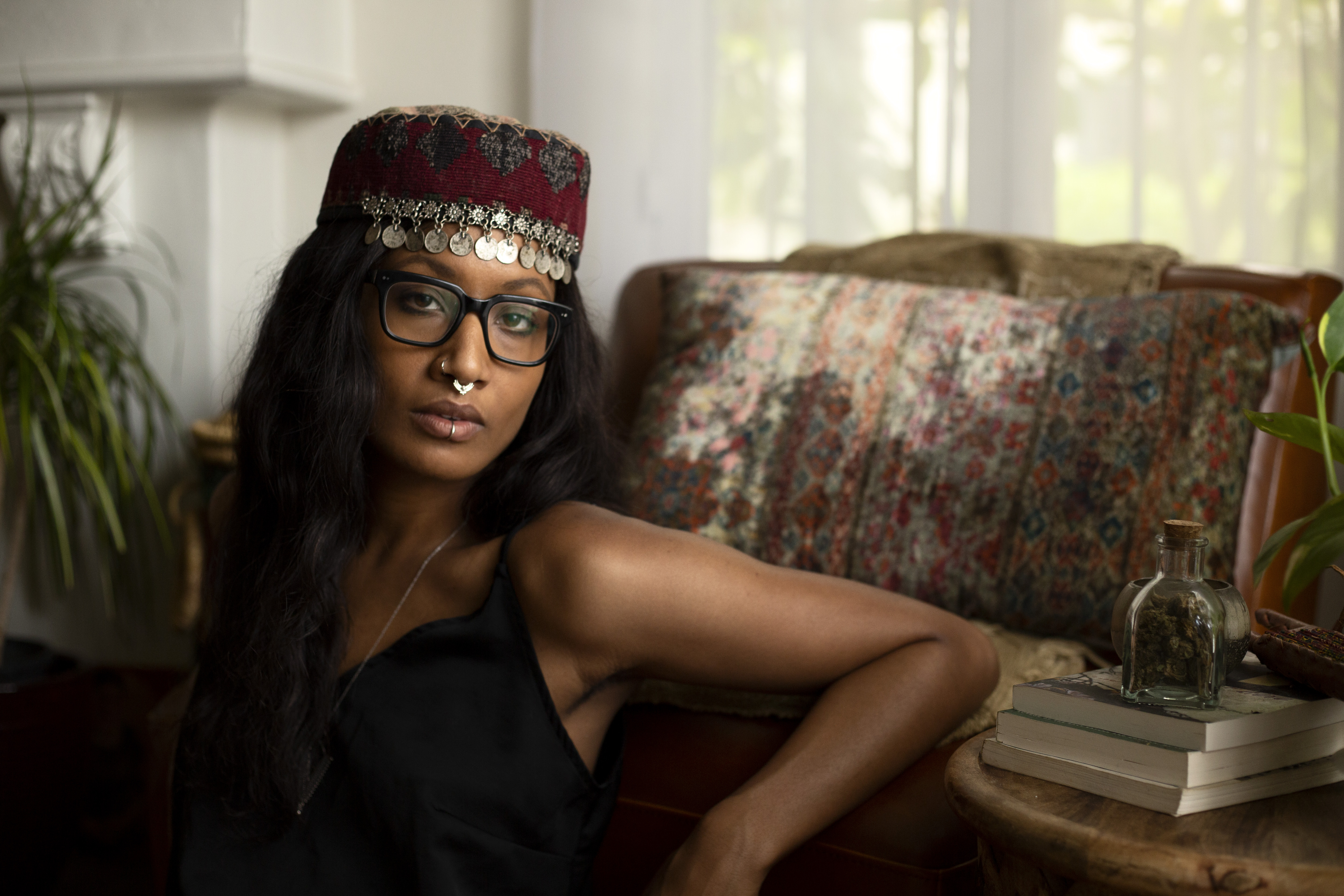 A Black woman with Glasses and a hat with jewels hanging over the forehead leans against a chair with her arm resting on it.