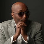 black bald man wearing sunglasses and a gray Suit
