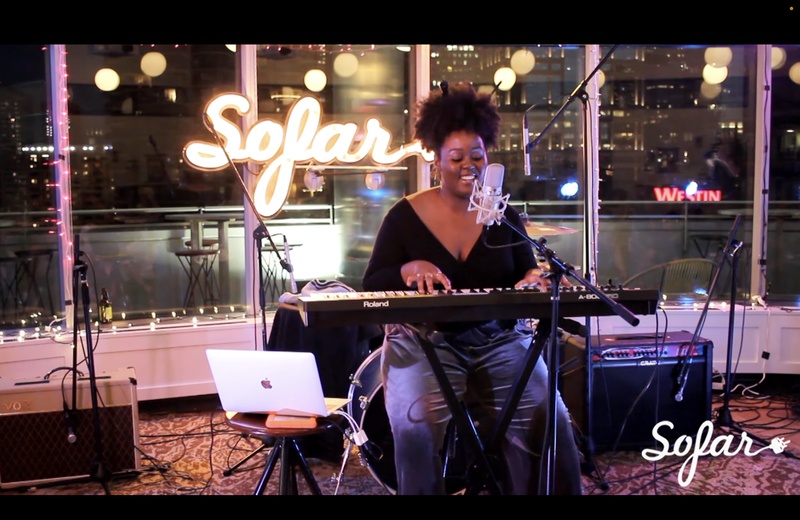  Black woman with curly hair tied up, sitting at a keyboard with a smile on her face in front of a neon sign that reads "Sofar" 