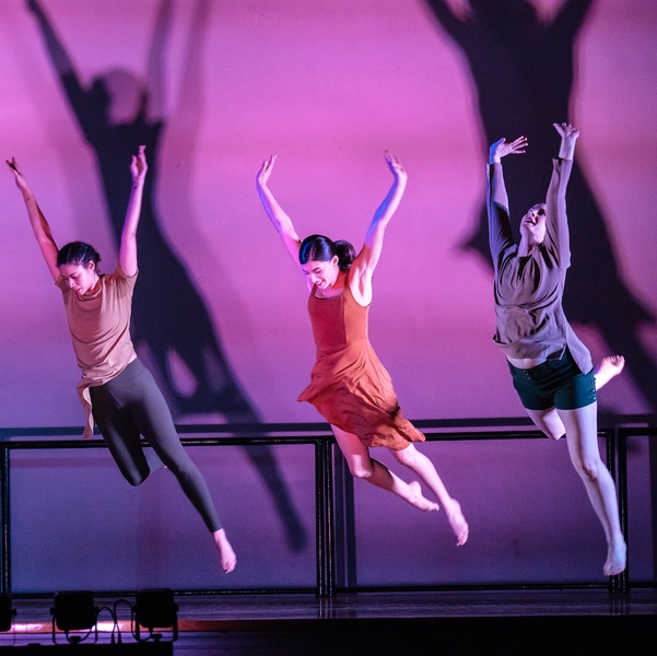  Three dancers in mid-air jumps with their hands stretched up above them and heads angled down to the floor. 