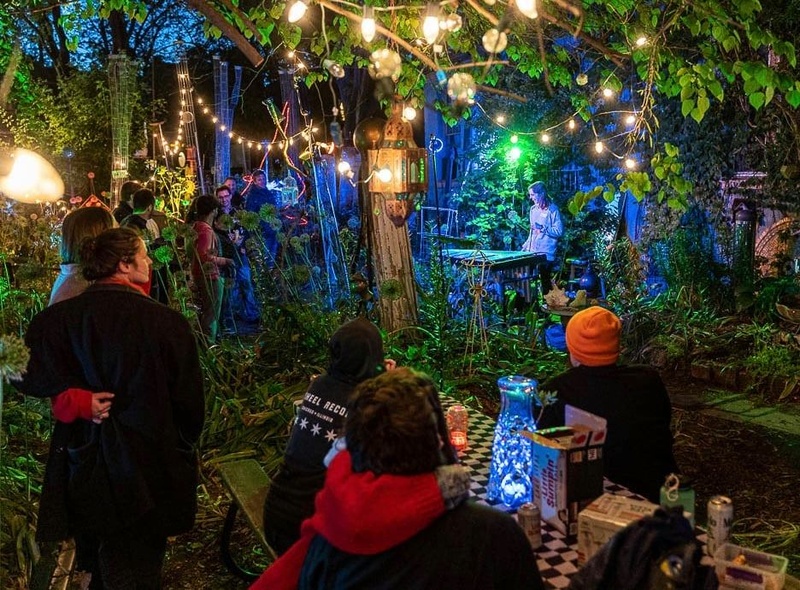  A white woman with wavy brown hair plays the vibraphone, from the center of a magical-looking garden full of happy spectators. The garden is full of vibrant greenery and decorated with many colored lights. 