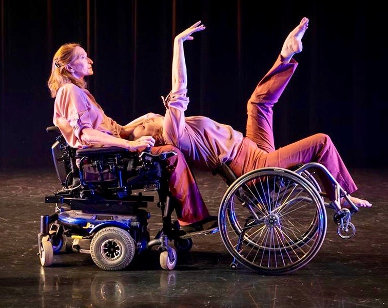  Ladonna tilts way back in her wheelchair with an arm and leg extended and upper body supported by an older female dancer using a motorized wheelchair 