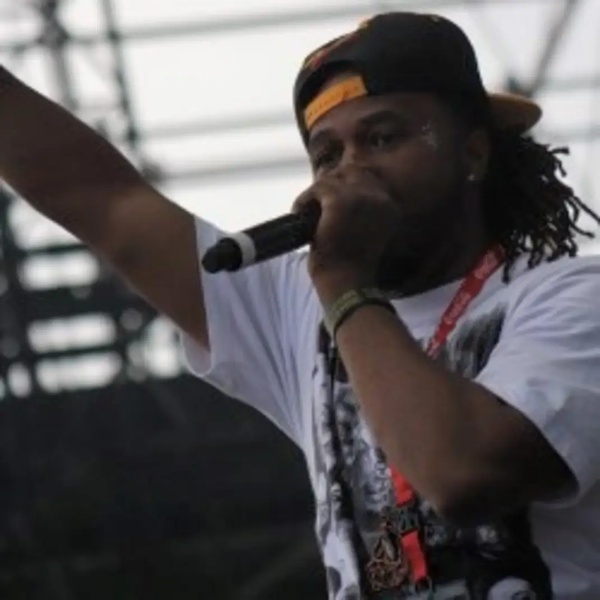  black man wearing hat points to the sky and speaks into microphone 