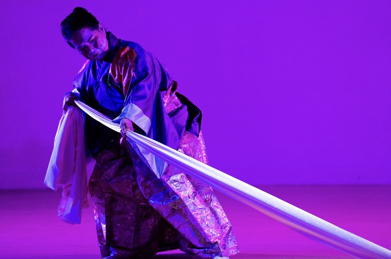  An Asian American woman dressed in traditional Japanese kimono pulling a long white fabric while cast in a purple light. 