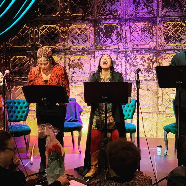  Two black women stand on stage in behind music stands and deliver a script reading 