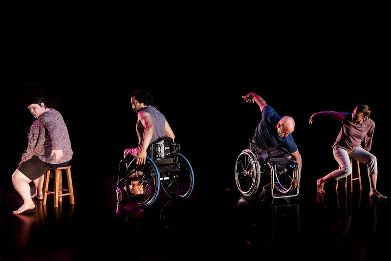  Four dancers move on stage. dancers on outside of group sit on stools, dancers on inside are in wheelchairs 