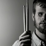 White man with beard and blue prosthetic eyes holds two drumsticks