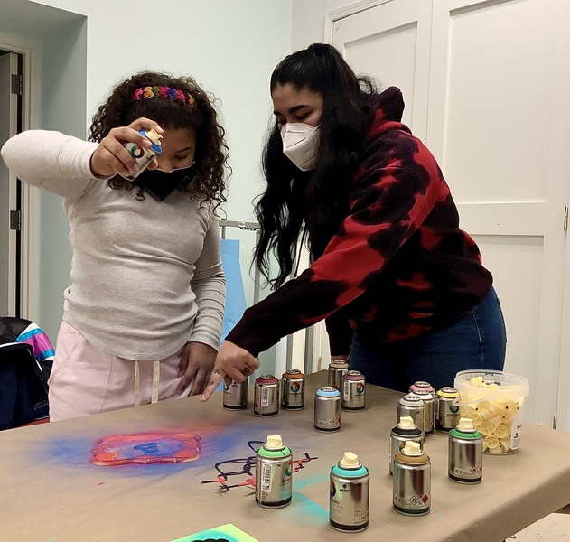  adult with long black hair and red/black tie die hoodie instructs small child on how to spray paint 