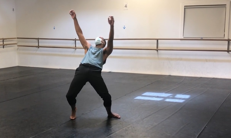  Black man with facemask in a dance studio, standing with knees gently bent as if almost falling backward, and his arms flung towards the ceiling. 