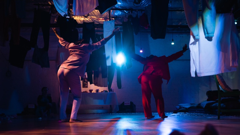  two dark skinned women standing with their backs to the camera one knee bend and their arms stretched up in a lavendar and red suit clothes hanging from the ceiling and a ligth from a video projected on the wall in dark lighting 