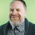 A middle aged white guy with dirty blonde hair, a red and grey beard smiles widely, almost uncomfortably. He wears his favorite teal sweater and a rather complex floral patten shirt. The photo is shot from the chest up  with a green background.