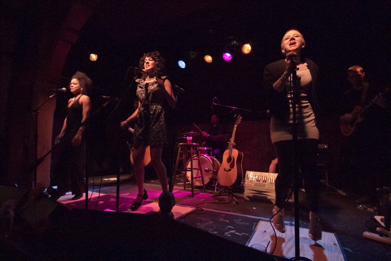  Ami sings and performs in the center of a dimly lit image. Stage lights light the dark stage with varying colors (blues, yellows, purples). Beside her, two performers sing. 