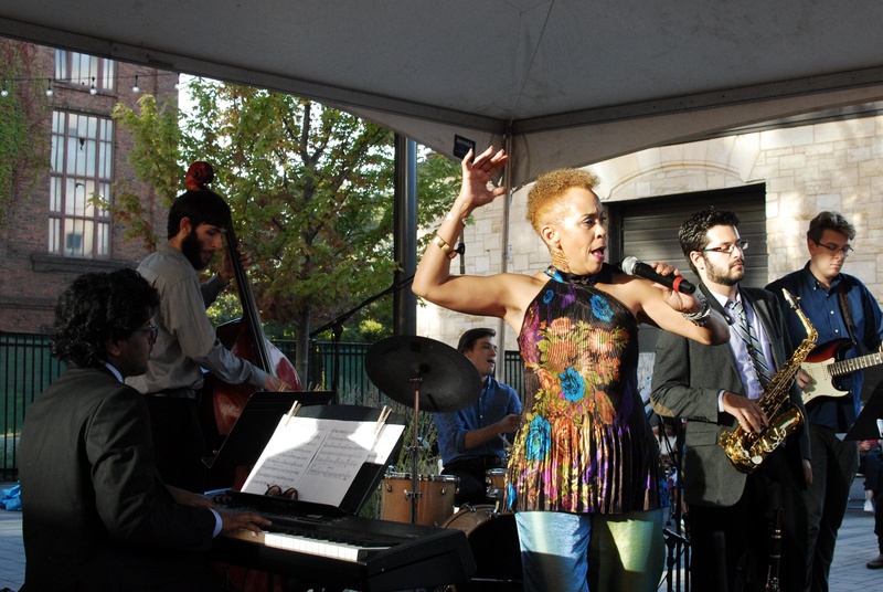  Woman with colorful top sings surrounded by a band, which includes piano player, drums, cello, guitar, and saxophone. 