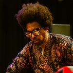 Black man with facial hair, large afro, mirrored sunglasses, a chain neckless, and a patterned shirt