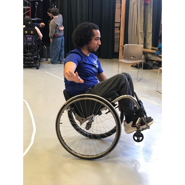  A multiracial Black and Mexican-American man with curly black hair balances on the back wheels of a wheelchair 