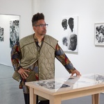 Black man short hair on the sides, long on the top,with glasses, with an oversized vest, standing in a gallery