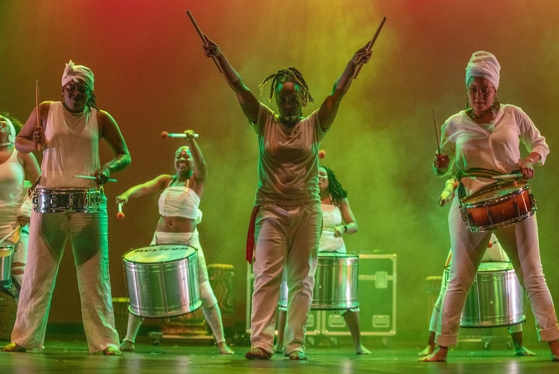  Six women wearing white stand on a stage playing drums slung around their waists, with colorful lighting in the background 