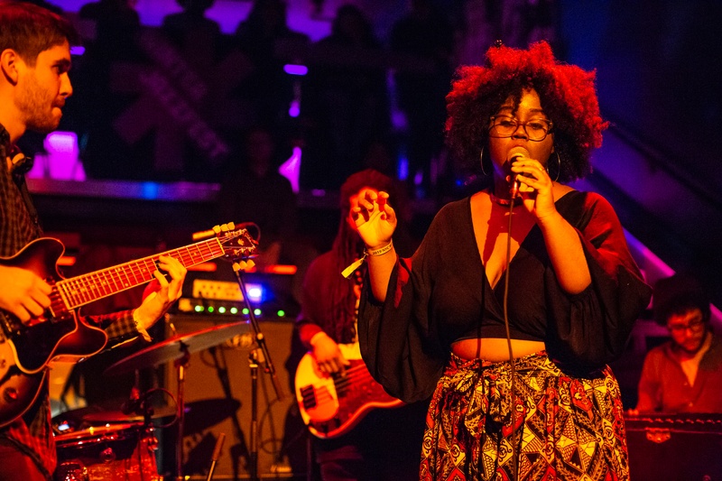  Black woman with curly hair and glasses holds a microphone on stage with other musicians. 