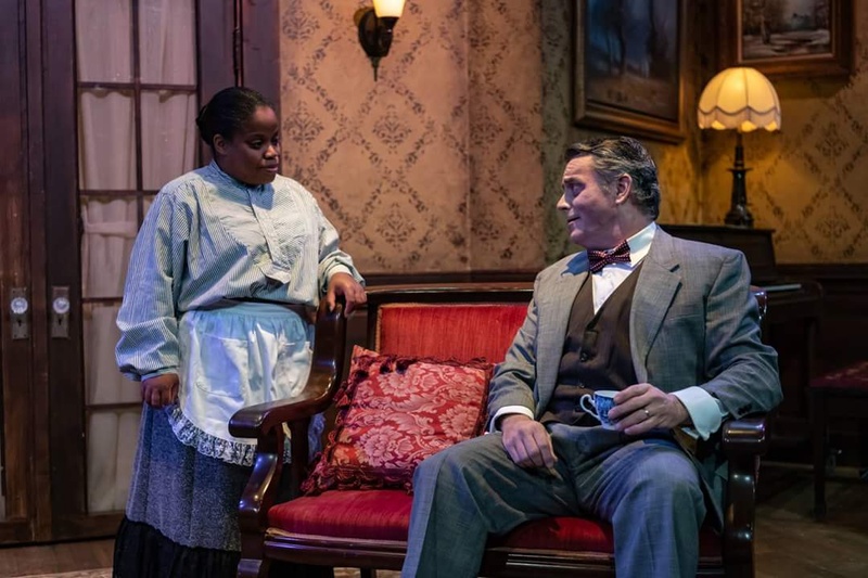  A Black woman on a stage set speaking to a person sitting on a red couch. 