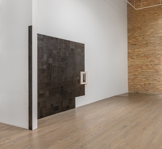  Angled view of a wall-area with a shimmery drawing made directly on the wall. At first, it appears black, then it looks metallic. The proportions correspond to the gallery's architectural features. A smaller framed work is situated towards the right. 