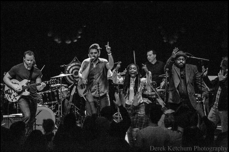  Black and white image of a six musicians assembled onstage in front of a boisterous and clapping crowd 