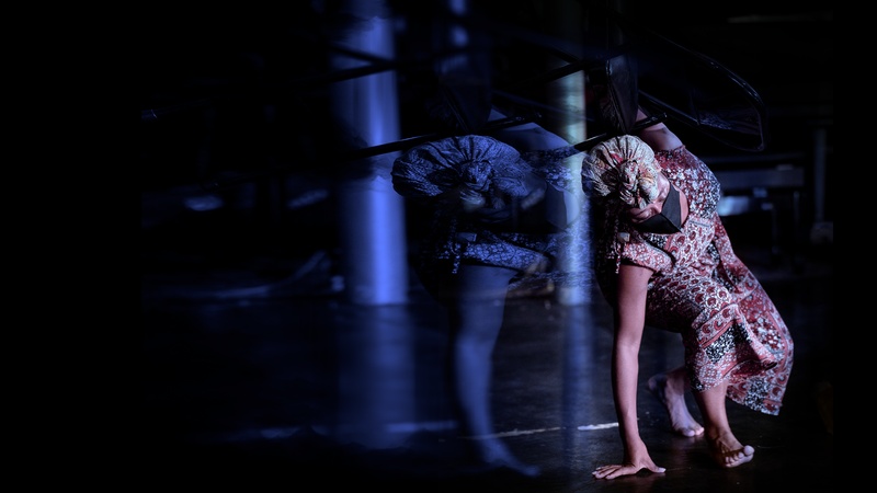  Black woman with patterned headwrap and dress, mask over her face, steps low and touches the ground with one arm. A softly lit reflection of her image is duplicated to the left, all against a darkly lit background. 
