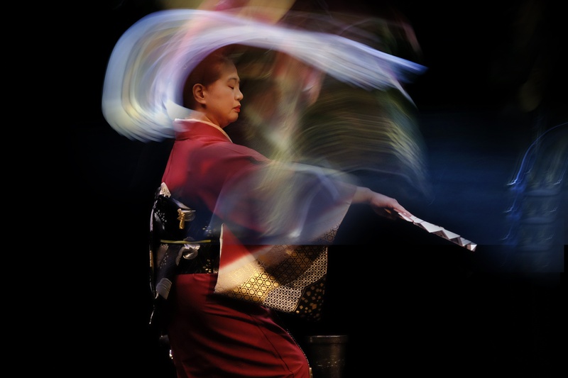  A Grand Master of Japanese traditional dance, Yoshinojo is standing erect with a fan in her outstretched hand, the spirits of her ancestors floating above her in a shimmering white cloud. 