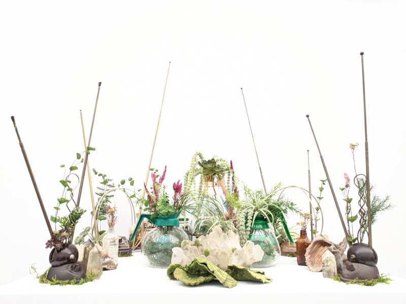  art installation on white pedestal with white background consisting of two vintage TV antennas, large quartz cluster, crystals, preserved dyed green polypore mushrooms,  two vintage restaurant coffee pots, dried/preserved plants and artificial plants 