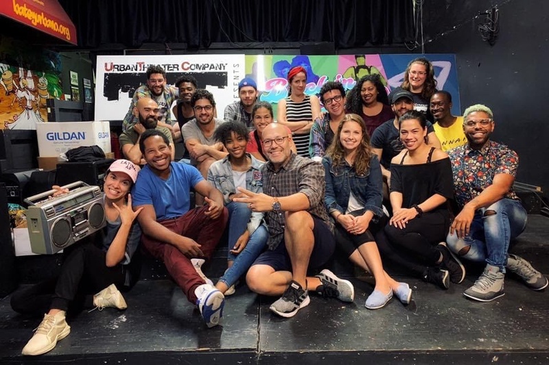  Group of 20 individuals in three rows smiling at the camera, seated or standing on a stage with Urban Theater Company signage in the background 