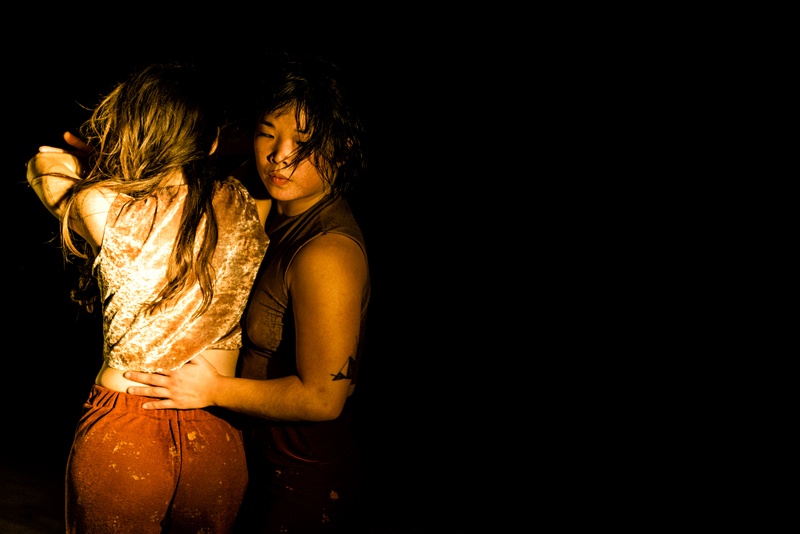  A White woman with long brown hair and an Asian woman with chin length hair pass through an emrace within a dark theater.  An amber glow hightlights their shimmering costumes and sweaty skin and hair. 
