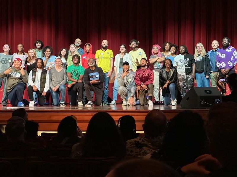  large group of students and teachers stand on stage in front of large crowd 