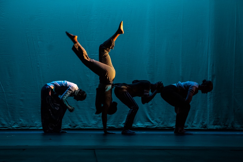  Four dancers in profile, side by side. Three are bent at the waist, heads down to the floor. The fourth does a handstand with legs raised at bent angles. 