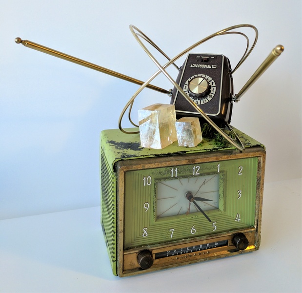  Vintage green and gold metal clock radio with a vintage TV antenna sitting on top of it, pointing out at viewer, translucent orange calcite crystals in square and rectangular shapes that appear to be leaning to the left, placed flat atop the clock radio. 