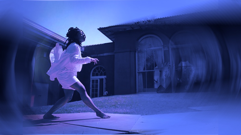  Black woman with long locs, wearing white shorts and tunic steps gracefully in profile on an outdoor stage. The left and right sides of the image are softly blurred like a camera focusing. 