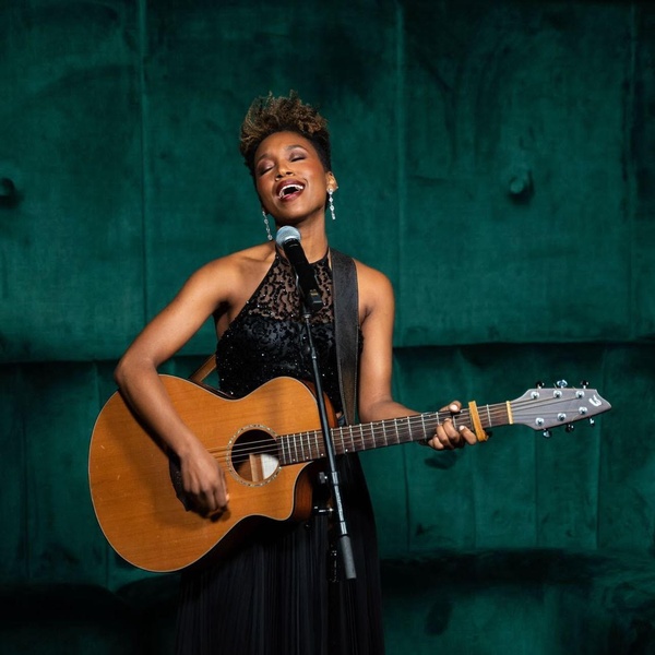  Black woman with short hair and blonde highlights stands with her guitar before a microphone, her mouth opened in song and eyes closed 