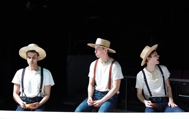  Three figures in straw hats, white shirts, and suspenders are assembled in a scene onstage 