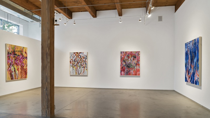  Four large and colorful paintings hang in a gallery space. The walls are white and the wooden ceiling and a beam are visible. 
