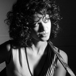 Ami has curly black hair and is photographed in black and white. She stares wistfully off to the side of the image and appears to be about to play an instrument.