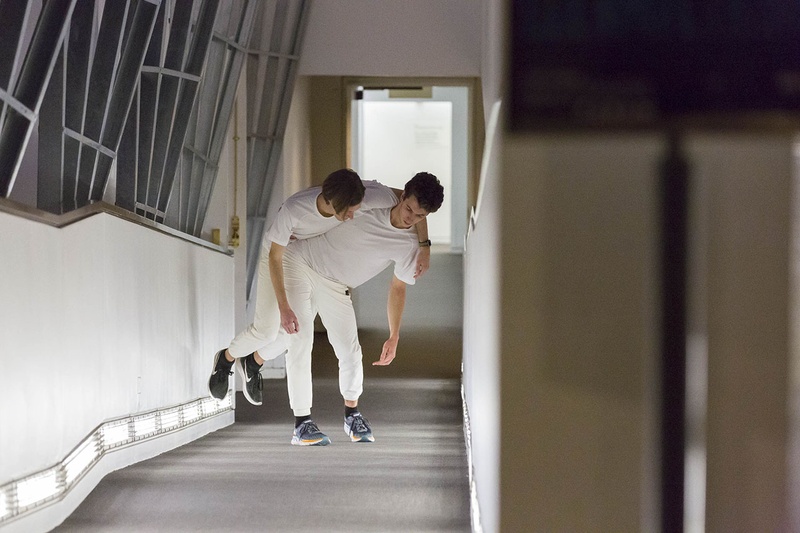  Two male dancers dressed all in white, one carrying the other on his back, slung over, walking up an incline in a narrow hallway 