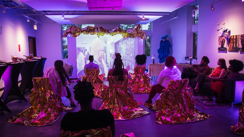  various people sitting in gold fabric colored seats facing a white wall with a video projection next to more people sitting on a greay couch with 3 picture of black women above them 