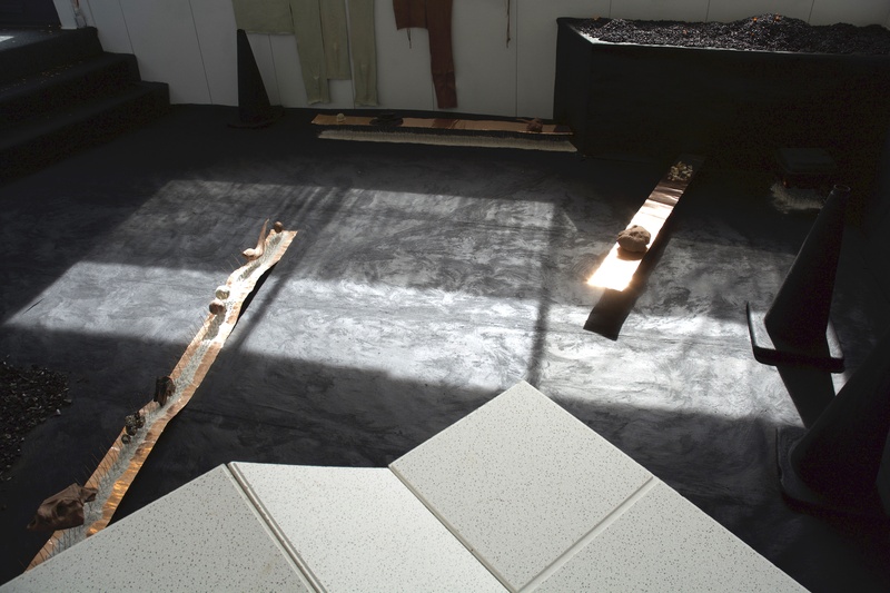  A minimalist room with an asphalt floor, traffic cones, and cascading white ceiling tiles. Copper foil stretches hold clay and organic objects, some on bird spikes. Sunlight filters in, casting shadows and illuminating the central area. 