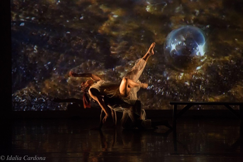  Black woman, softly lit onstage, seemingly suspended in the air with arms and legs raised up and away from her body as if gently falling or rising, with a dark celestial sky and silver orb in the background 