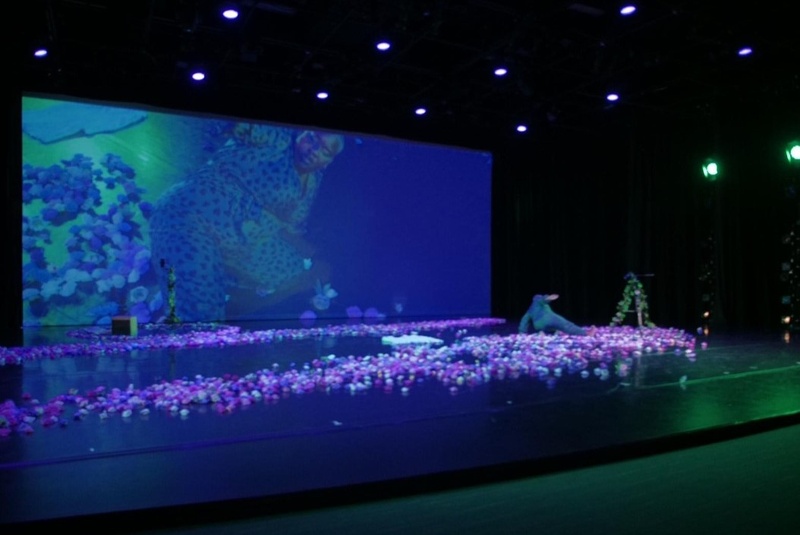  black dark skinned woman in dark green jumpsuit knealing down on a bed of pink, whit, and purple flowers, with a black camera on a tripod covered in green vines dancing in frontof a big screen projecting her image on the bach screen with purple lighting 