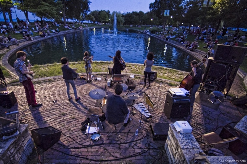  Ami performers outside surrounded by band members. In front of them is a small body of water and a listening crowd. 